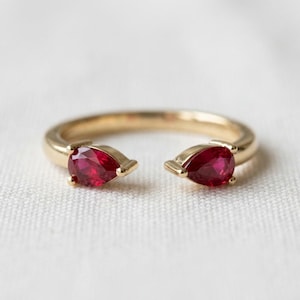 May include: A gold open-style ring featuring two pear-shaped, deep red rubies set at each end of the band. The band is smooth and polished, with the two gemstones facing each other across the open gap, creating a balanced and symmetrical design.