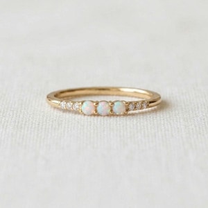 May include: A gold ring featuring three round, iridescent opals set in the centre, flanked by a series of small, clear, round-cut stones along the band. The metal band has a polished finish and a thin, delicate profile.