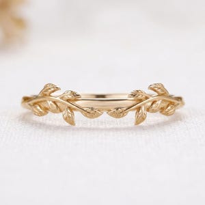 May include: A gold ring featuring a delicate band adorned with two symmetrical, sculpted leaf motifs that extend from the center toward the sides. The metal has a polished, reflective finish, highlighting the detailed texture of the leaves.