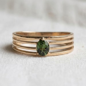 May include: A gold-toned ring featuring a multi-band design with four parallel horizontal bars. An oval-cut, deep olive green gemstone is set centrally, connecting the four bands. The metal has a polished finish and a warm, metallic lustre.