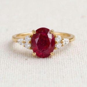May include: A gold ring featuring a large, oval-cut deep red gemstone at the center. The band is accented on both sides by a cluster of three small, clear, round-cut white stones, creating a symmetrical design with a polished metal finish.