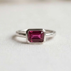 May include: A silver-toned ring featuring a rectangular, emerald-cut deep pink gemstone set in a smooth bezel setting. The band is polished and solid, with a clean, minimalist design that highlights the vibrant color of the central stone.