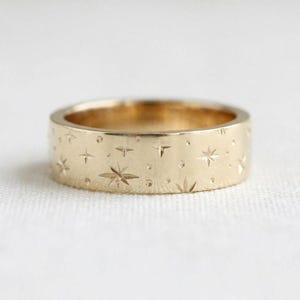 Peut inclure: Une bague anneau en or avec une surface plate gravée d'un motif répétitif de petites étoiles à plusieurs branches et de minuscules points circulaires. Le métal présente une finition lisse et polie, avec des motifs d'étoiles répartis uniformément.