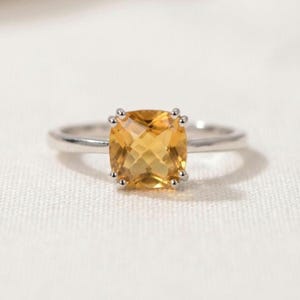 Könnte beinhalten: Ein silberfarbener Ring mit einem quadratisch geschliffenen, facettierten gelben Citrin, der in einer Vier-Krappen-Fassung gehalten wird. Die Ringschiene ist glatt und poliert und zeigt ein klares, metallisches Finish, das den warmen Goldton ergänzt.