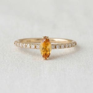 Puede incluir: Un anillo de oro con una piedra preciosa de citrino amarillo en talla marquesa sujeta por cuatro garras. La banda delgada está acentuada con una hilera de pequeños diamantes blancos redondos y claros engastados a ambos lados de la piedra central.