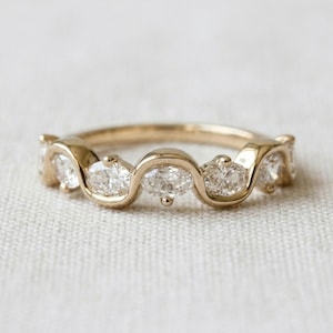 May include: A gold ring featuring a continuous wavy band design. The ring is set with five oval-cut clear white diamonds arranged in a row, with each stone held within the curved gold metal sections of the band, creating a fluid and cohesive look.