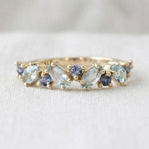 May include: A gold ring featuring a series of alternating marquise-cut light blue stones and round-cut dark blue stones set along the band. The metal band has a polished finish and includes the engraved text ALE on the inner surface.