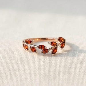 May include: A rose gold ring featuring a delicate vine-like band adorned with marquise-cut orange-red gemstones arranged in a leaf pattern. The metal band has a polished finish, and the stones are set securely along the top half of the ring.