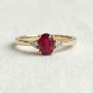 Könnte beinhalten: Ein Goldring mit einem zentralen, oval geschliffenen, tiefroten Rubin in einer Vier-Krappen-Fassung. Die Ringschiene besteht aus poliertem Gelbgold und ist auf jeder Seite des zentralen Rubins mit einem kleinen, rund geschliffenen weißen Diamanten besetzt.