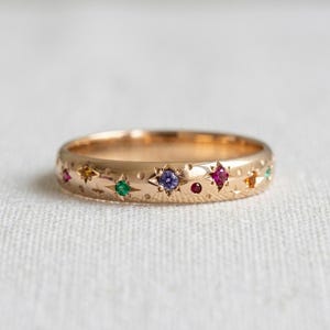 Peut inclure: Une bague en or avec une série de pierres précieuses multicolores serties à fleur, notamment des pierres violettes, rouges, vertes et jaunes. La surface métallique est ornée de motifs d'étoiles gravés et de petites indentations circulaires.