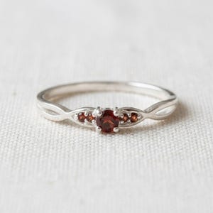 Könnte beinhalten: Ein Silberring mit einem zentralen, rund geschliffenen, dunkelroten Granat-Edelstein, der auf jeder Seite von zwei kleineren, rund geschliffenen Granatsteinen flankiert wird. Die Ringschiene hat ein geteiltes Design, das zur Fassung hin zusammenläuft.