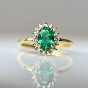 May include: A gold ring with an oval emerald gemstone at its centre, encircled by a halo of small, round diamonds. The ring's band is plain, polished gold. The emerald is a vivid green, and the diamonds sparkle against the gold setting.