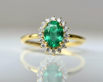 May include: A gold ring featuring an oval-cut emerald center stone surrounded by a halo of small, round diamonds. The ring's band is a simple, polished gold. The emerald is a vibrant green color, and the diamonds sparkle against the gold.