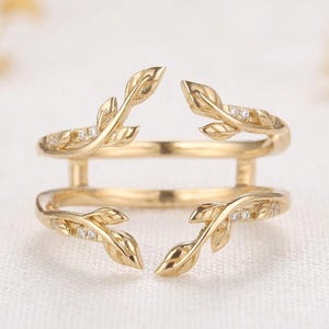 May include: A gold open-style ring featuring two parallel bands connected by vertical bars. Each band terminates in a delicate leaf motif, with small clear stones embedded along the surface of the leaves, creating a nature-inspired design.