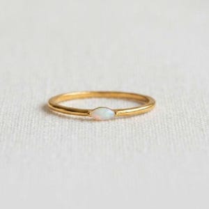 Könnte beinhalten: Ein dünner Goldring mit einem einzelnen, kleinen, marquiseförmigen Opal, der horizontal in der Mitte gefasst ist. Das Metall hat eine polierte Oberfläche und der schillernde Stein zeigt subtile Lichtreflexe in Weiß, Blau und Rosa.