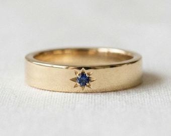 May include: A gold band ring featuring a single round blue sapphire set in the center. The gemstone is encased within a starburst-shaped engraving, creating a focal point on the smooth, polished surface of the yellow gold metal band.