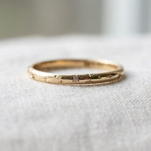 Peut inclure: Une fine bague en or avec une finition polie et des motifs d'étoiles gravés répartis uniformément sur la circonférence. Une petite pierre précieuse ronde et transparente est sertie au centre de l'un des motifs d'étoiles gravés sur la bague.
