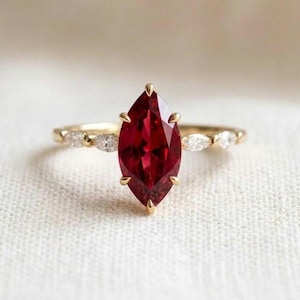 May include: A gold ring featuring a large marquise-cut deep red gemstone at the centre, held by four prongs. The band is accented on each side by three small, clear marquise-cut stones set in gold, creating a delicate and symmetrical design.