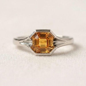 May include: A silver-toned ring featuring a square-cut, golden-yellow gemstone set in a bezel setting. The band has a distinct architectural design with three parallel grooves extending from the central stone along each side of the metal shank.