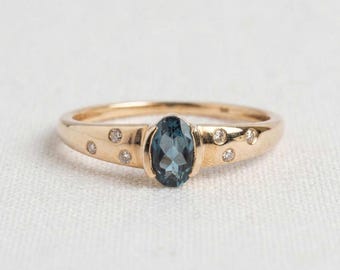 May include: A gold ring featuring a central oval-cut blue gemstone held in a bezel setting. The band is accented with six small, round, clear stones embedded into the metal on either side of the center stone, creating a symmetrical design.