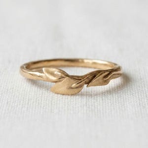 Könnte beinhalten: Ein goldener Ring mit einem zarten Band und einem skulpturalen Design aus zwei überlappenden Blättern in der Mitte. Das Metall hat ein weiches, mattes Finish und die Blätter sind mit feinen Adern und spitzen Enden geformt, was eine organische Form ergibt.