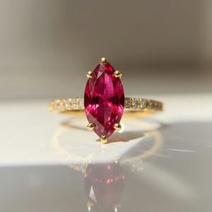 May include: A gold ring featuring a marquise-cut, deep red gemstone. The band is adorned with small, sparkling stones. The ring is set against a bright, reflective surface, highlighting its elegant design and vibrant color.