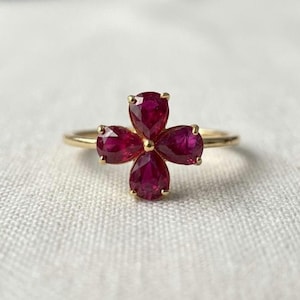 May include: A gold ring featuring a flower-shaped cluster of four pear-cut deep red rubies. The stones are arranged symmetrically around a central point, held in place by gold prongs on a thin, polished gold band that encircles the finger.