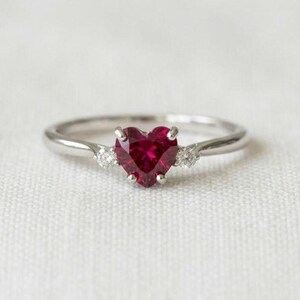 Op de afbeelding: Een zilverkleurige ring met een centraal hartvormige rode edelsteen in een zetting. De band is aan weerszijden van het hart voorzien van twee kleine, ronde, heldere stenen, wat zorgt voor een symmetrisch ontwerp met een gepolijste afwerking.