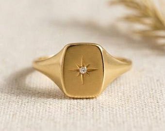 May include: A gold signet ring featuring a rectangular face with rounded corners. The center of the ring face is engraved with a starburst pattern containing a single small, clear, round-cut gemstone at its core, set against a smooth finish.