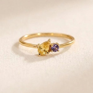 May include: A thin gold ring featuring two small gemstones set side by side. One stone is a yellow pear-shaped gem, and the other is a smaller, round purple gem. The band is smooth and polished, showcasing a delicate design with these two distinct coloured stones.