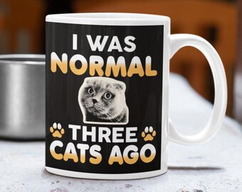 May include: White ceramic mug with a black background featuring the text "I WAS NORMAL THREE CATS AGO" in yellow and white. A cat's face is in the center. The mug has a white handle.