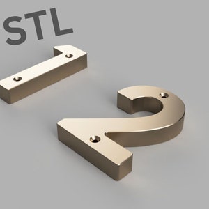 Może przedstawiać: Dwa złote metalowe numery domów, 1 i 2, z płaskim tyłem i wypukłym przodem. Numery są przeznaczone do montażu na ścianie lub powierzchni.