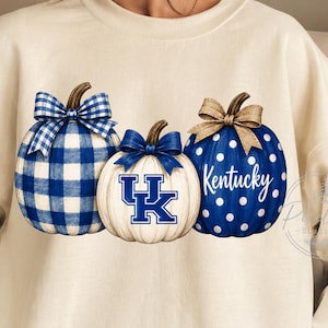 May include: A cream-colored crewneck sweatshirt featuring a graphic of three pumpkins. The left pumpkin has a blue and white buffalo check pattern with a blue bow. The center white pumpkin displays a blue UK logo. The right blue pumpkin has white polka dots, a tan bow, and the word Kentucky.