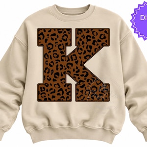 May include: A beige crewneck sweatshirt featuring a large, block-style letter K centred on the front. The letter is filled with a brown and black leopard print pattern. A purple circular badge in the top right corner contains the text Digital File.
