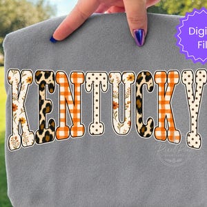 May include: A grey t-shirt featuring the word KENTUCKY in large block letters. Each letter has a unique pattern including orange and white gingham, leopard print, brown polka dots, and a floral design with orange flowers and green leaves.