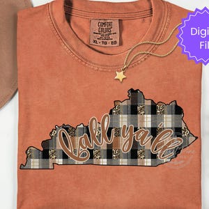 May include: A terracotta-colored t-shirt featuring a graphic of the state of Kentucky filled with a black, white, and brown plaid and leopard print pattern. The text 'y'all' is written in a cursive brown font across the center of the state shape.