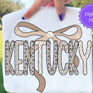 May include: A white t-shirt featuring the word KENTUCKY in a bold, leopard print pattern. Behind the text is a large, light brown ribbon bow with a central white pearl accent. A purple circular badge in the corner contains the text Digital File.
