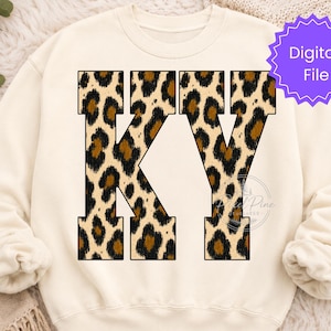 Peut inclure: Un sweat-shirt à col rond de couleur crème arborant les lettres KY dans une police de caractères grasse et en bloc. Les lettres sont remplies d'un motif imprimé léopard marron et noir. Un badge circulaire violet contient le texte Digital File.