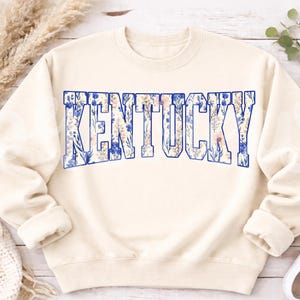 May include: Cream-colored sweatshirt with the word "KENTUCKY" in large, blue-outlined letters. The letters are filled with a floral pattern in shades of blue, white, and pink. The sweatshirt has long sleeves and a crew neck.