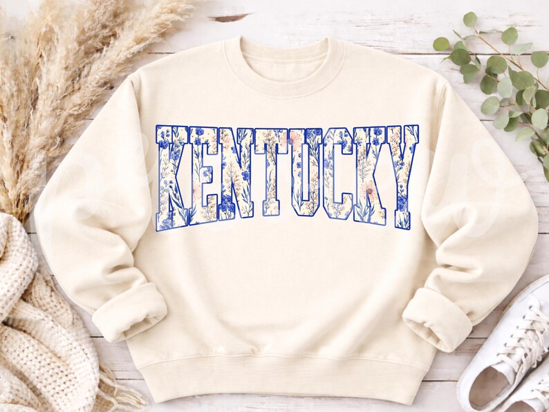 May include: Cream-colored sweatshirt with the word "KENTUCKY" in large, blue-outlined letters. The letters are filled with a floral pattern in shades of blue, white, and pink. The sweatshirt has long sleeves and a crew neck.