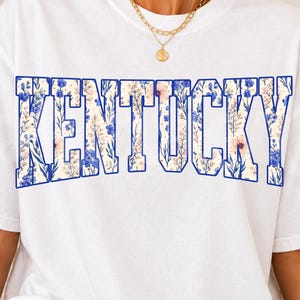 May include: White t-shirt featuring the word "KENTUCKY" in large, block letters. The letters are filled with a floral pattern in shades of blue, pink, and cream, outlined in blue. A gold necklace is visible at the top.