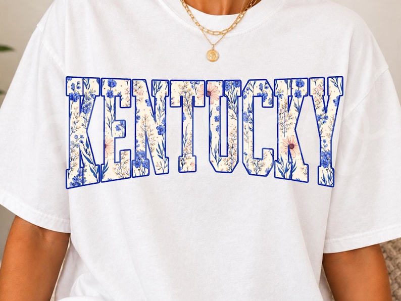 May include: White t-shirt featuring the word "KENTUCKY" in large, block letters. The letters are filled with a floral pattern in shades of blue, pink, and cream, outlined in blue. A gold necklace is visible at the top.
