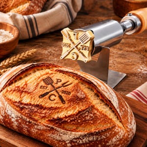 May include: A metal branding iron with a wooden handle, featuring a brass head with a crossed spatula and fork design, the letters P and C, and the text EST 2020. The branding iron is positioned above a loaf of bread with the same design branded on it.
