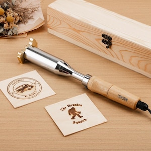 May include: A wood-burning tool with a wooden handle and a metal tip, alongside two engraved wooden squares. One square features a car graphic, the other a Bigfoot design. A wooden box is in the background.