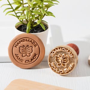 Puede incluir: Un sello redondo marrón con las palabras "HANDMADE JANE CLACK" y un diseño de mariposa. También es visible un sello dorado con un mango de madera y el mismo diseño. Una pequeña planta en maceta está en el fondo.