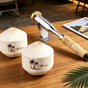 May include: Two white coconuts with a palm tree graphic and the name Catherine printed on the side, positioned next to a silver electric branding iron with a wooden handle and a metal stand. The branding iron features the text 110V/300W.