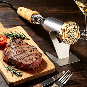 May include: An electric branding iron with a brass head featuring the text MADE BY FARAZ ALI EAT MY MEAT and a steak icon. The tool has a wooden handle and rests on a metal stand next to a seared steak on a wooden board with rosemary and tomato.