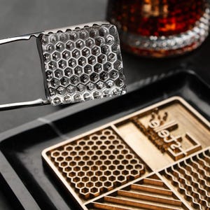 Op de afbeelding: Een heldere, vierkante ijsblokje met een honingraatpatroon, vastgehouden door een roestvrijstalen tang. De ijsblokje bevindt zich boven een houten dienblad met een honingraatpatroon. Een glas met een donkere vloeistof staat op de achtergrond.