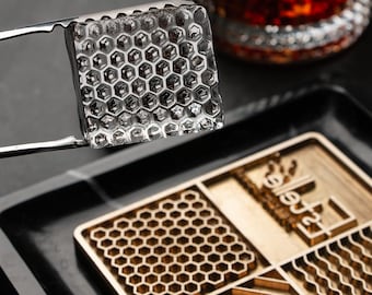 May include: A clear, square ice cube with a honeycomb design, held by stainless steel tongs. The ice cube is above a wooden tray with a honeycomb pattern. A glass with a dark liquid is in the background.