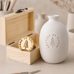 May include: A white ceramic vase with a narrow neck and a matte finish, featuring the embossed text POTTERY BY KAREN and a pine tree icon. Beside it sits an open light-coloured wooden box containing a circular brass stamp with a pine tree design.